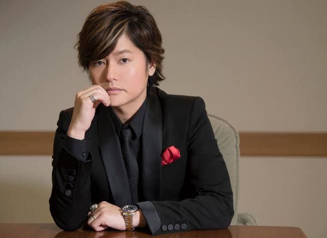 Showtaro Morikubo สวมสูทสีดำพร้อมผ้าเช็ดหน้าสีแดง นั่งเท้าคางอยู่ที่โต๊ะไม้